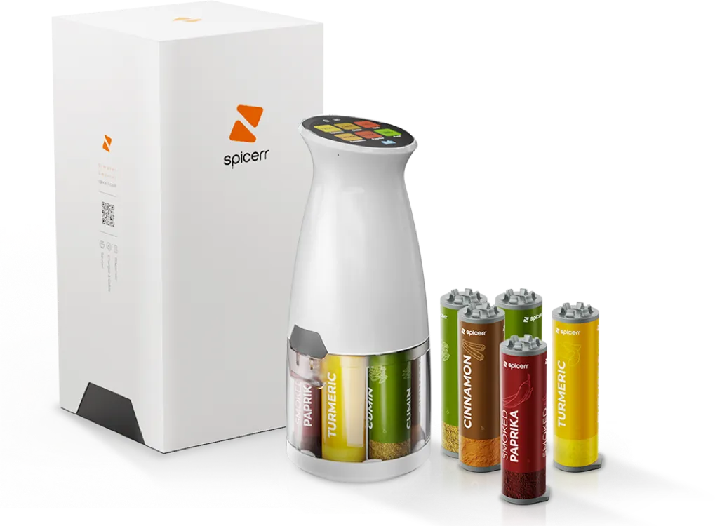 A white automated spice dispenser with multiple spice cartridges labeled turmeric, cumin, cinnamon, and smoked paprika is shown next to packaging.