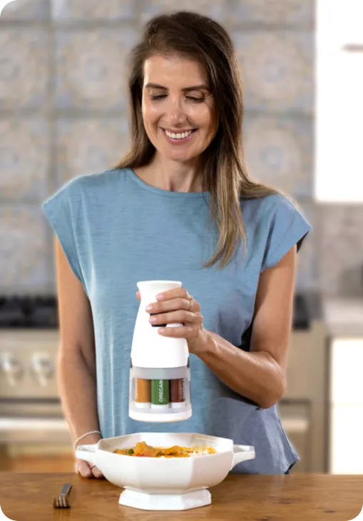 A person in a kitchen grinds spices with a white grinder over a bowl. The background features patterned tiles and a stove.