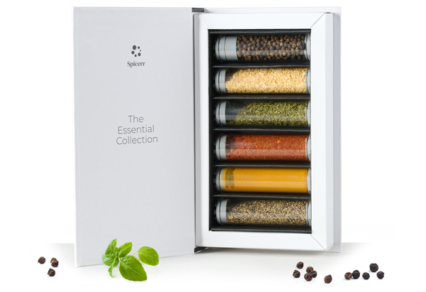 Open box containing six spice vials labeled "The Essential Collection." Includes scattered peppercorns and fresh basil nearby on a white surface.