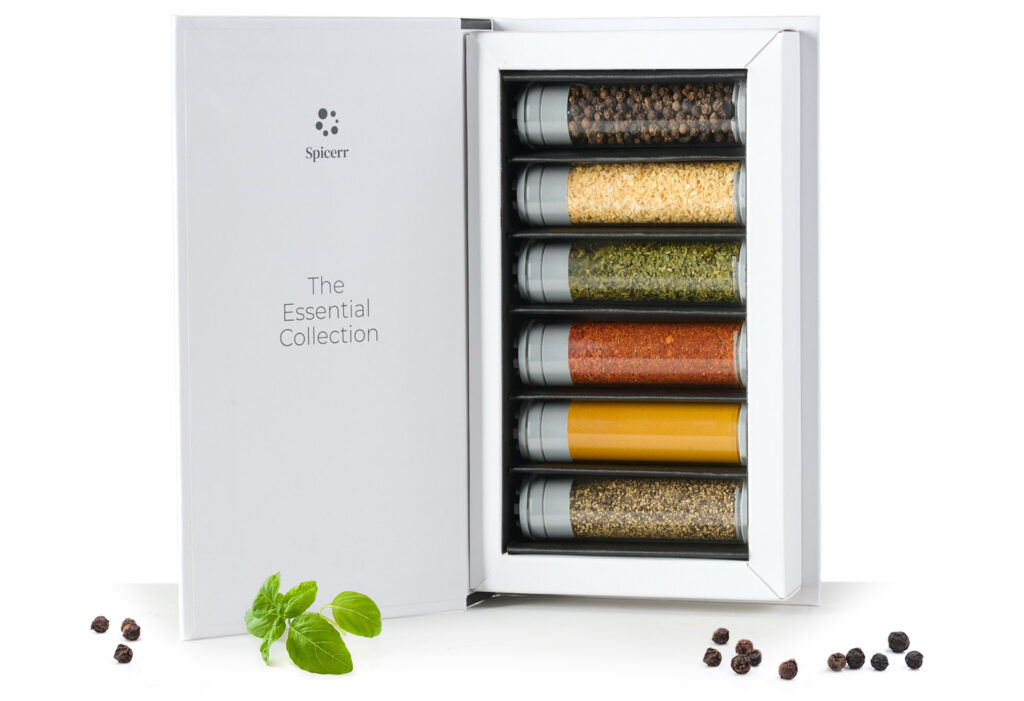 A white box labeled "The Essential Collection" contains six spice jars. Basil leaves and scattered peppercorns are placed nearby for decoration.